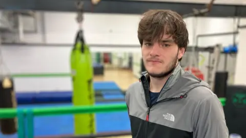 BBC / Kate McGough Niall looks into the camera. He has medium-length brown hair down over his forehead and is wearing a grey North Face zipped-up jacket. In the blurred background we can see he is in a boxing gym.