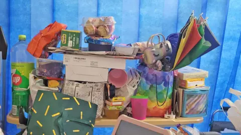 Jessie's brightly coloured possessions - including pink cups, a rainbow umbrella, art materials, a glasses case, as well as drinks and snacks, are piled on the small table next to her hospital bed. The blue curtain is pulled around it for privacy. 