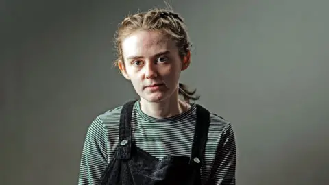 Stephanie stands against a grey backdrop and looks directly at the camera. She has her hair in plaits and is wearing blue dungarees and a stripey jumper.