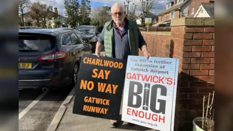 Emily Coady-Stemp/BBC Peter Barclay the chairman of the Gatwick Area Conservation Campaign with two large placards stating opposition to the expansion of the Airport