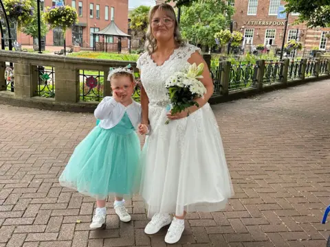 Claire Claire and Emilia pictured on Claire's wedding day. Claire is wearing a white, tea-length wedding dress with a lace v neck top half and a diamante belt, with an a-line tulle skirt. She has white trainers and is holding white flowers. Emilia is in a turquoise bridesmaid dress with a tulle skirt, white bolero cardigan and white trainers. They are pictured outside on a paved area with gardens behind them.