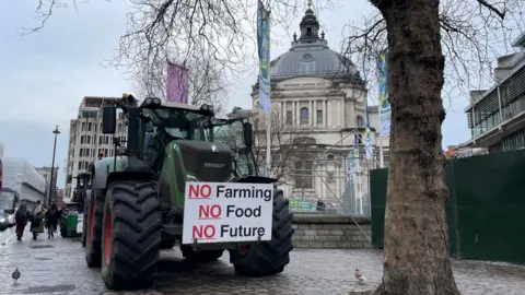 Malcolm Prior/BBC Tractors parked outside conference centre in a wet London street. The lead tractor has a placard which says 