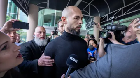 Reuters Andrew Tate, bearded and wearing a black sweater, is surrounded by press outside of an airport terminal