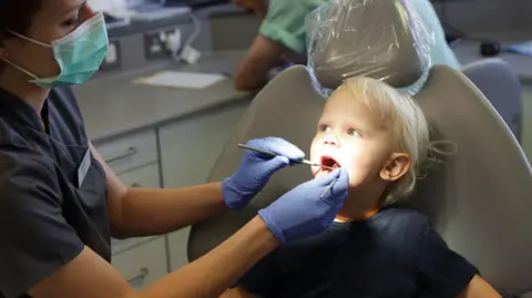 Getty Images A young blonde boy sat in a dentist chair. He is looking at a female dentist who is looking in his mouth with two dental tools.