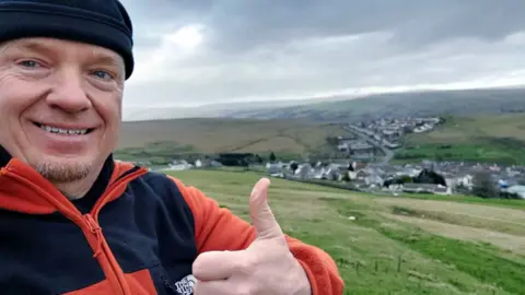 Instagram | Robert Charles Albon Joe Albon looks towards the camera in the middle of a field, with green countryside behind him. He is wearing a black hat, and a black and orange jumper. He is holding his thumb up and smiling at the camera.