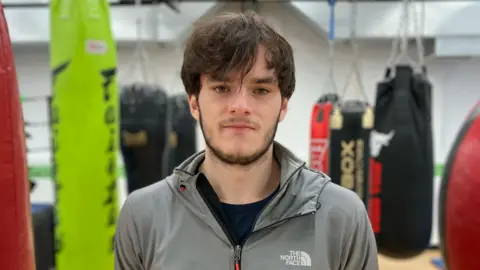 BBC /Dan Nelson Niall looks into the camera. He has medium-length brown hair down over his forehead and is wearing a grey North Face zipped-up jacket. In the blurred background we can see he is in a boxing gym.