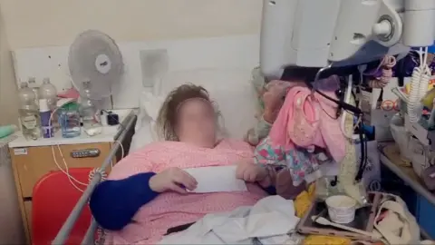 BBC Jessie wearing a pink top and holding a letter lies in her hospital bed, surrounded by all her possessions, some of which are hanging from the monitor. 