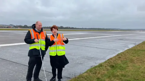 BBC News Bronwen Jones, Development Director at London Gatwick standing on the northern runway at Gatwick airport alongside BBC transport correspondent Sean Dilley, pointing to a grass verge where the runway will be expanded under the airport's proposals. Both are wearing high vis jackets.