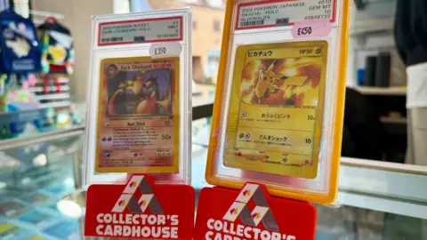 Danny Fullbrook/BBC Two Pokemon cards stood upright on a counter in Collector's Cardhouse branded displays. One is a Dark Charizard card, the other is Pikachu card with Japanese text. 