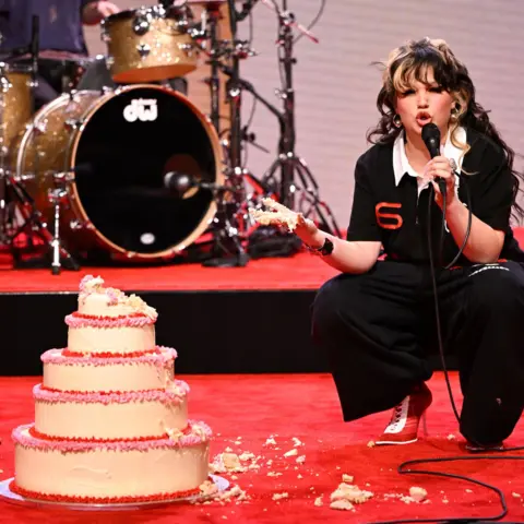 Getty Images Lola Young performing on the Jimmy Fallon show. She wears black trousers and a black polo shirt and crouches next to a five-tiered cake with cream, red and pink frosting. She's gouged out the top tier and her cake is covered in cake. She's performing on a red carpet and behind her on set is a golden drum kit.