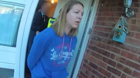 BBC Lucy Letby wearing a blue hooded sweater coming out of her house in handcuffs with a police officer following her. She is looking past the camera, and is wearing her blonde/brown shoulder length hair down.