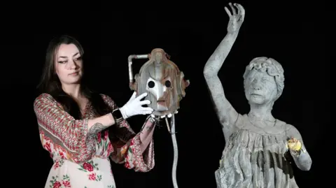 PA Media A woman is holding a decapitated Cyberman head next to a weeping Angel statue which is missing wins and a left hand.