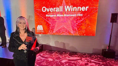 Slimming World Mrs Miles-Bramwell holding a trophy, and behind her is a screen saying 