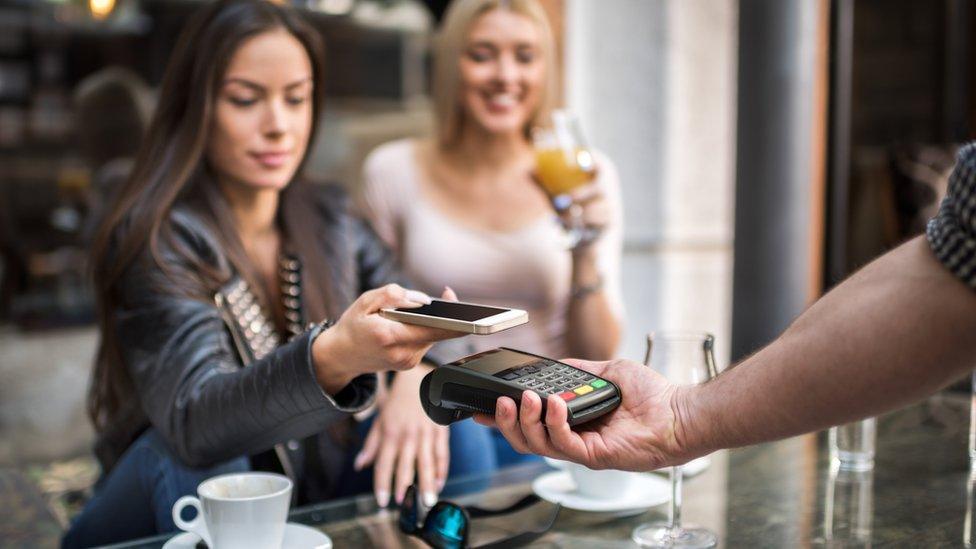 A smiling woman with long dark hair and wearing a leather jacket uses her mobile phone to pay for a coffee in a café. Another woman with blond hair and wearing a white t-shirt is drinking a glass of orange juice.