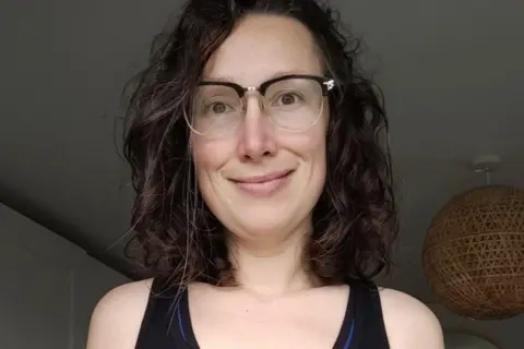 Alix Harvey A woman with dark hair, a black top and glasses smiles at the camera. She is stood in a house