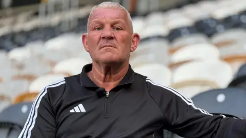 BBC Newsnight Windass, with short grey hair, is sitting on a black seat in Hull City's football stadium. He is looking directly into the camera lens and wearing a black sports top with white stripes down the side.