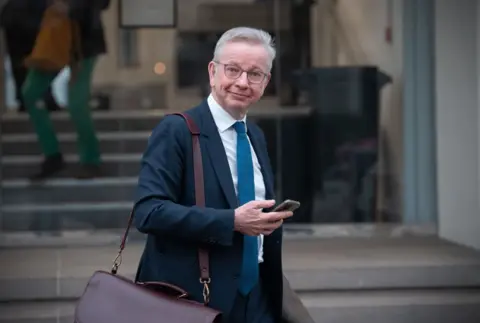 Getty Images Michael Gove standing outside the Covid Inquiry on Monday, 10 March, after giving evidence. He is wearing a dark blue suit and staring straight at the camera while checking his mobile phone.