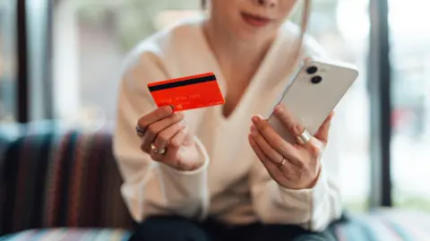 Getty Images Midsection of young Asian woman holding a credit card while using smartphone, sitting on sofa. Online shopping and electronic payment.