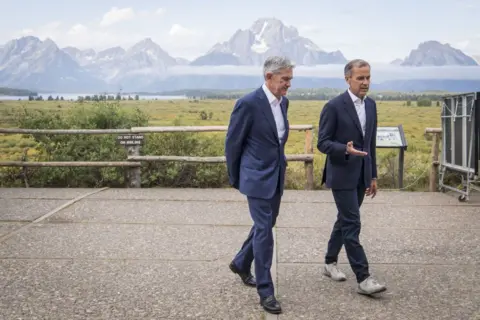 Getty Images Jerome Powell, chairman of the US Federal Reserve (left) and Mark Carney, former governor of the Bank of England, walk the grounds of Jackson Hole economic symposium in 2019. Both wearing blue suits, they are surrounded by lush green fields with the mountains in the background