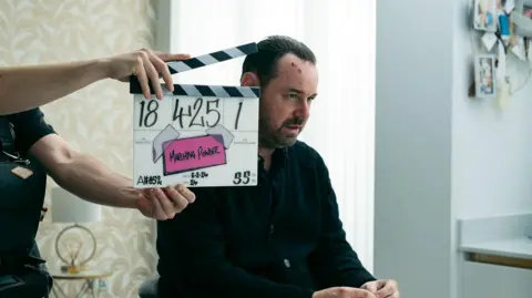 TRUE BRIT Danny Dyer looks ahead of him with blood on his forehead, dressed in black and sitting down. He has a serious expression, a clapper board to his left and arms holding it. There are blinds behind his head and wallpaper to his left with yellow leaves on a white backdrop. To his right are photos hung up over a counter.