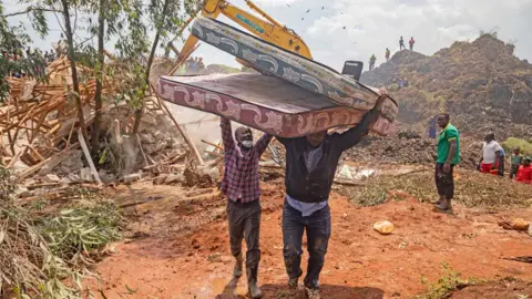 AFP A man holds up his hands to help another man carrying tow mattresses away from Kiteezi waste site after its collapse in August 2024 - Kampala, Uganda