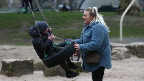 Getty Images A smiling woman with blonde hair wears as light blue coat as she pushes a grinning young boy on a tyre swing in a park. 