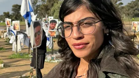 Oren Rosenfeld / BBC Michal Ohana, with long dark hair, large glasses and wearing pink lipstick, stands in front of a memorial to those who died in the 7 October attack on the Nova festival, with photos of the people killed, Israeli flags, flowers and other mementoes.