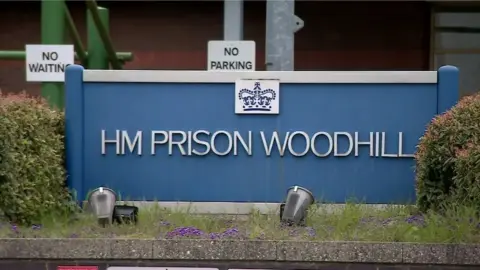 The sign outside HMP Woodhill - a large blue sign between two hedges. There are NO WAITING and NO PARKING signs behind it