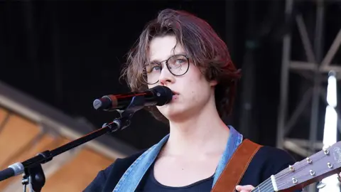 Getty Images David Kushner with a microphone in front of him and holding a guitar, wearing glasses 