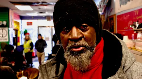 Darren Conway/BBC John White, a black man with a greying beard, who wears a dark woollen hat, a large grey coat and a red fleece underneath, photographed close-up at the drop-in centre with other attendees or staff visible out-of-focus behind him. A tear is rolling down his cheek.