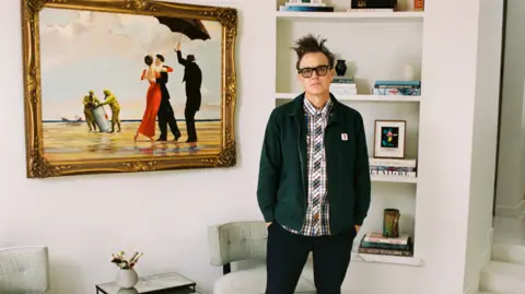 SOTHEBY'S Mark Hoppus wearing a green jacket over a checked shirt and dark jeans. He is standing in front of a white wall and bookcase. The painting is on the wall to his right.