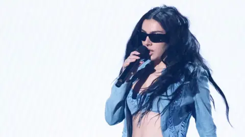 Reuters Charli XCX in a denim jacket and bra-top, sings into a microphone during the 2025 Grammy Awards