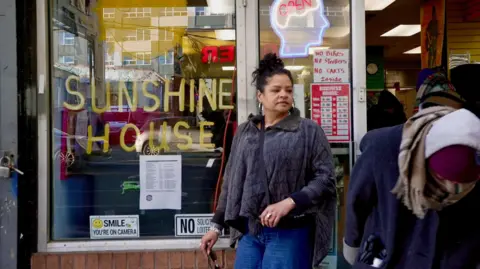 Darren Conway/BBC Rosalind Pichardo, a woman with her black her up in a bun, wearing a grey top and blue jeans, standing outside her drop-in centre which has 