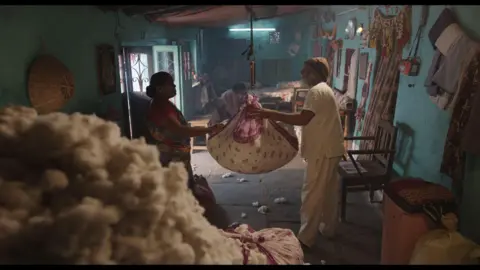 Courtesy: Sthal A still from Sthal: A Match shows an elderly couple weighing cotton on a scale hung from the ceiling inside a room. In the foreground is a huge pile of freshly picked cotton