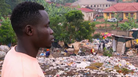 BBC Okuku Prince in a pink T-shirt looking out over the Kiteezi rubbish dump in Kampala, Uganda, with houses seen in the background.