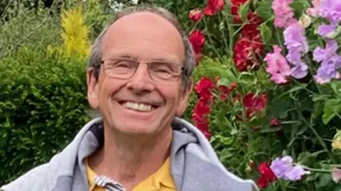 Stephen Stephen, who has short grey hair, back from a receding hairline, is wearing glasses and is smiling broadly. He has a grey sweatshirt over a yellow top and is standing in front of a hedge with red, pink and mauve sweet peas growing on his left.