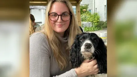 Holly Crouch Holly Crouch, a young woman with long, wavy blonde hair and tortoiseshell glasses, is wearing a grey jumper with a high neck as she holds her dog, a black and white spaniel, and smiles at the camera.