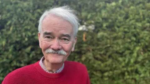 Peter Cunningham has grey hair and and grey moustache and is wearing a red round neck sweater. He is smiling.