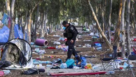 EPA An Israeli police officer walks through the Nova festival site after the attack, where, among the slender trees providing shade, tents are scattered around and personal possessions litter the floor after people fled. On one tree, a sign says, 