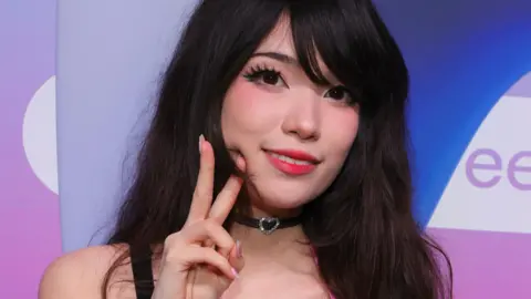 Getty Images Emiru at TwitchCon 2024. She has long dark hair worn loose and wears false eyelashes, red lipstick and blush. She makes a peace sign while smiling at the camera. 