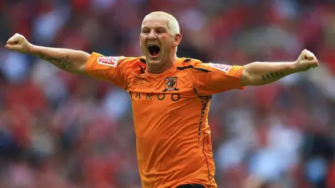 Getty Images Windass has short bleached blonde hair and is running off with both arms out wide on the pitch at Wembley. He is wearing Hull City's orange shirt.