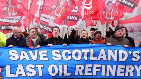 PA Media Trade unionists holding placards stand behind a blue banner which reads Save Scotland's last oil refinery