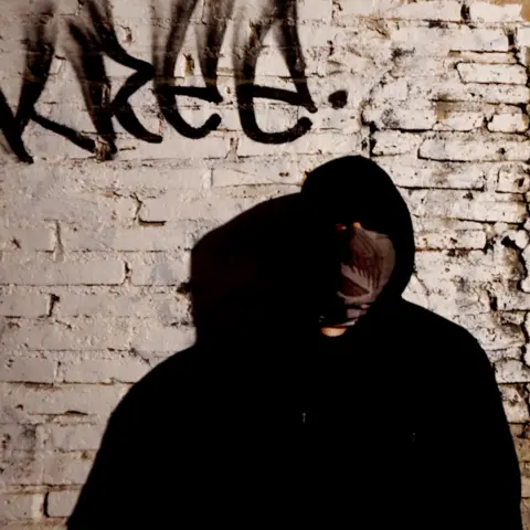 BBC A man standing in shadow and further concealed by a black hoodie and a bandana over his face, in front of the wall of a cartel safe house, which has crumbling brick and fading white paint. There is unintelligible graffiti above his head.