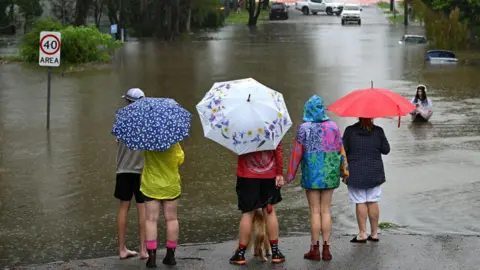 Getty Images Residents in the Brisbane suburb of Newmarket carry umbrellas and stand on dry ground next to a flooded part of the road