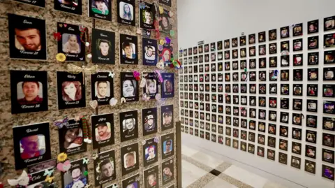 Darren Conway/BBC Two walls which are covered with the names and images of people who have died, tightly packed so there are 10 pictures in each column - and the columns continue beyond the edge of this photograph. Below each picture is the age the person died at: 