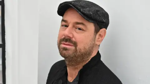 TRUE BRIT Headshot of Danny Dyer looking at the camera in a black cap, you can see the top of a black cardigan and black shirt. He has a warm expression in his eyes, mouth is neutral. There is a white wall behind him. 