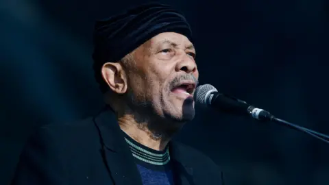 BBC Roy Ayers performing at Glastonbury in 2019. He has a blue top and black jacket on and is singing into the microphone on stage.