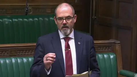 Parliament TV Paul Foster, in a dark blue suit and with receding grey hair and a grey goatee beard, speaks to the House of Commons with the green benches in the backdrop