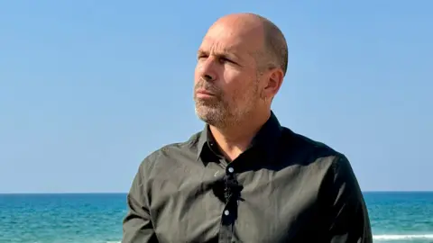 Oren Rosenfeld / BBC Roy Salomon, a man with a shaved head and stubble, with an earring in his left ear and wearing a dark buttoned-up shirt, standing by the sea with his back to the water