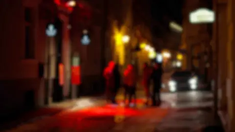 Blurry image, which has a red tint to it, of 5 men stood down alleyway 